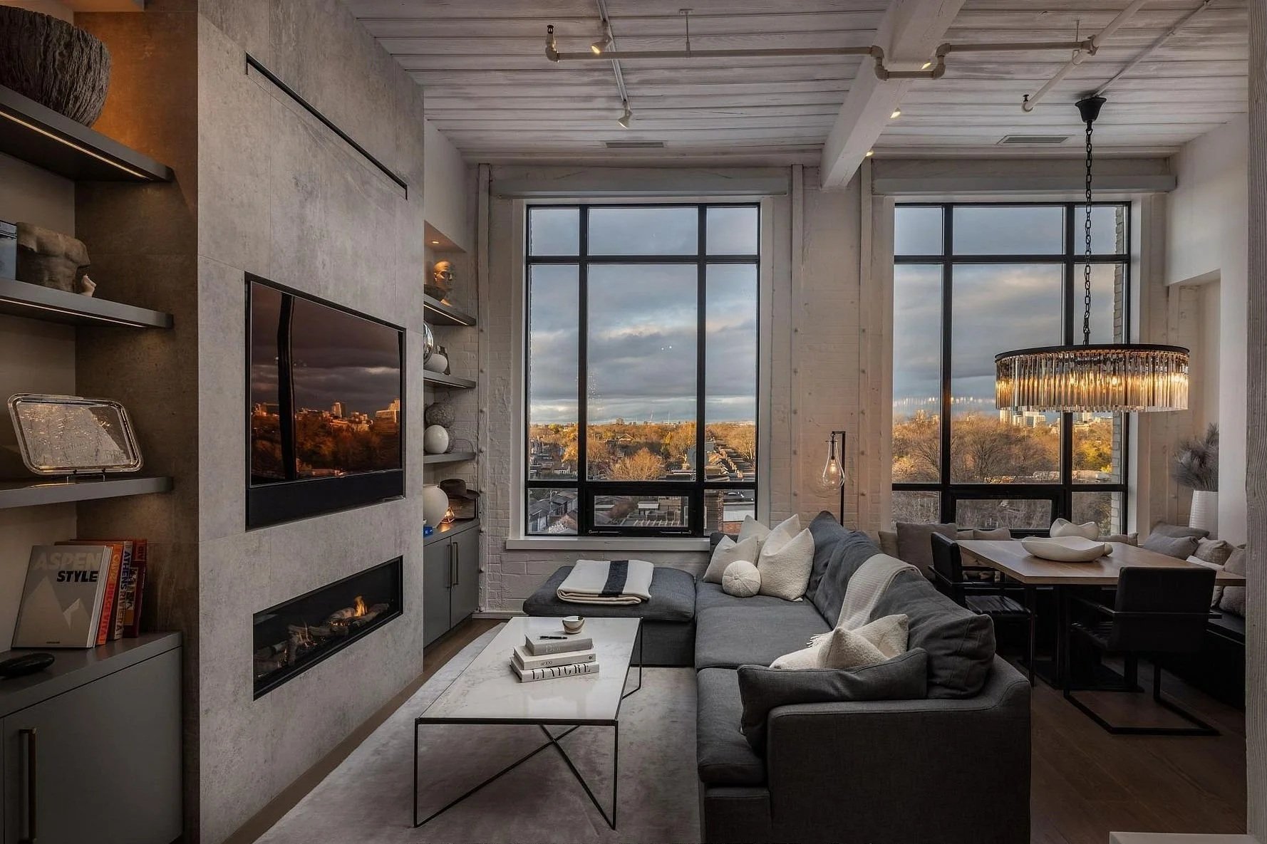 Modern living room with large windows showing a cityscape and trees, gray sectional sofa with white and gray pillows, coffee table with books and decorative items, built-in shelves, flat-screen TV, hanging chandelier, and dining area with a white table and black chairs.